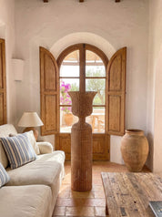 A rustic, bohemian-style living room featuring a tall, woven brown vase in the center of a tiled floor, flanked by a large clay pot, a cream sofa with striped pillows, and an arched wooden window with open shutters.