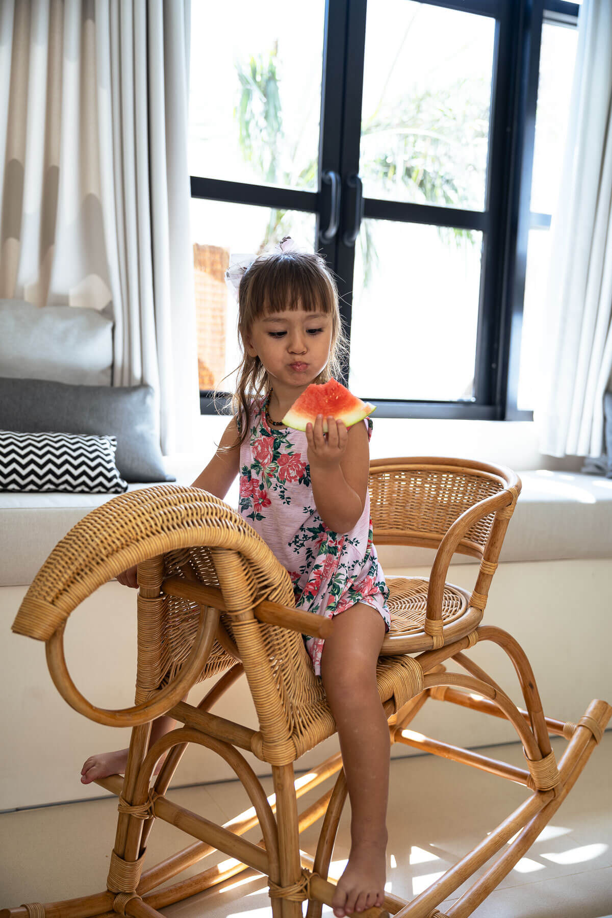 Child sitting on a wicker chair eating an orange in a bright room with large windows.