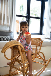 Child sitting on a wicker chair eating an orange in a bright room with large windows.