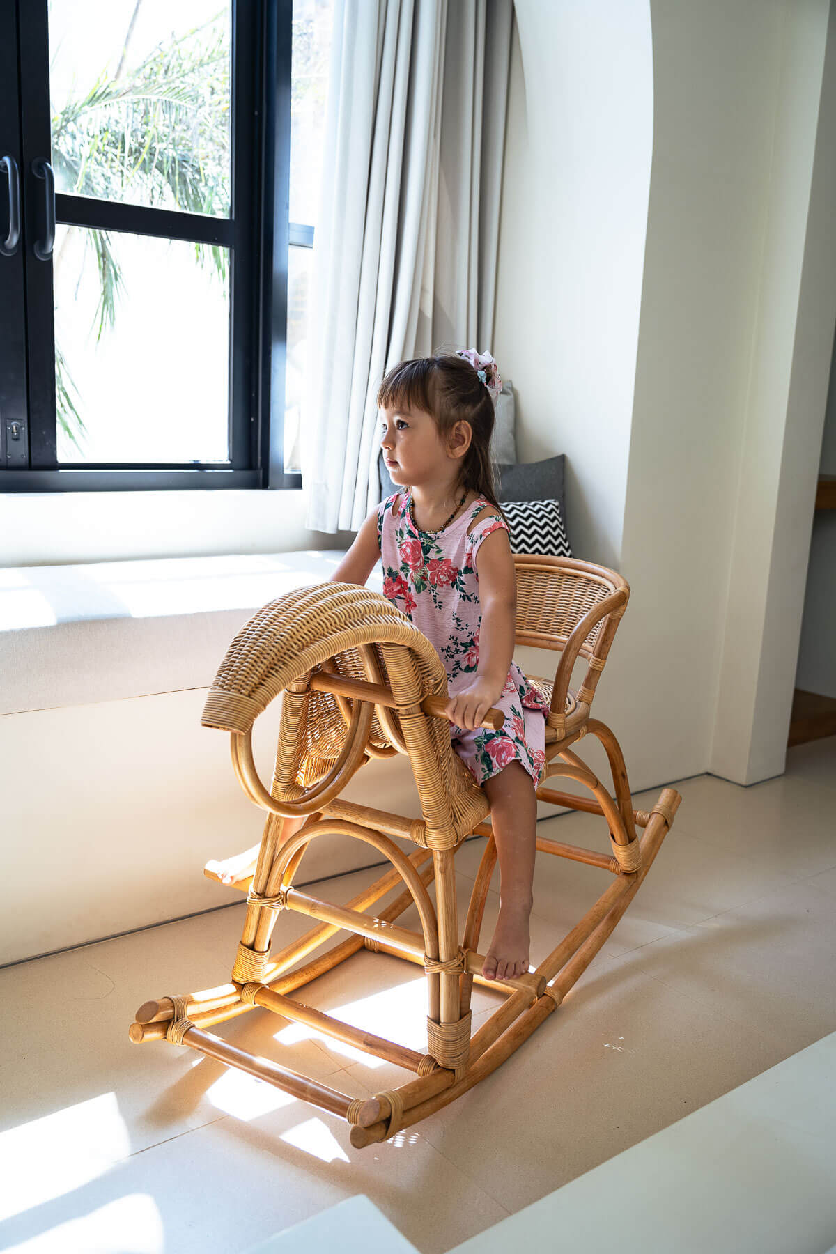 Child sitting on a wicker rocking horse in a bright room with large windows.