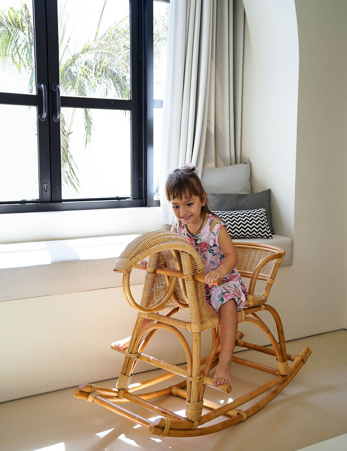 Child sitting on a wooden rocking chair in a bright room with large windows.