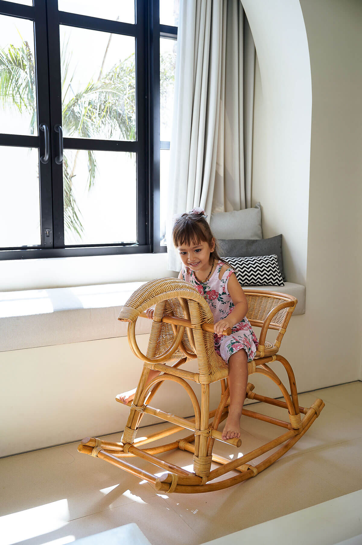 Child sitting on a wooden rocking chair in a bright room with large windows.