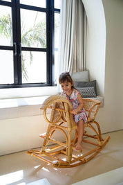 Child sitting on a wooden rocking chair in a bright room with large windows.