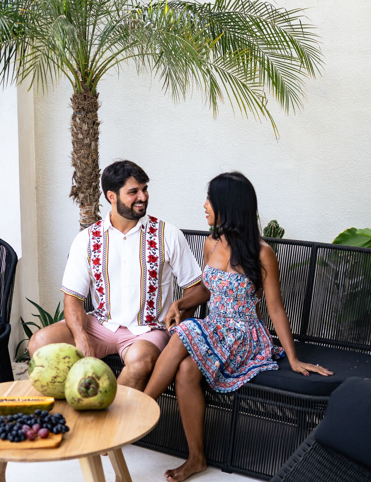 Two people sitting on a couch in an outdoor setting with a palm tree and white wall.