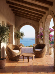 Two woven round outdoor armchairs with white cushions and a small round wooden coffee table on a terracotta tiled patio, set under stone arches with a view of the blue sea in the background.