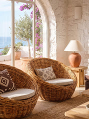 Two round woven wicker lounge chairs with cream cushions in a sunlit Mediterranean-style room, with an arched window showing the sea and a stone lamp on a side table.