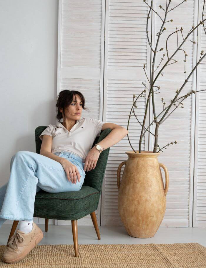 Woman sitting in a green chair next to a vase with branches indoors.