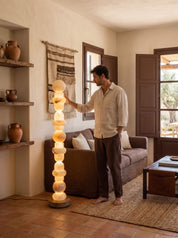 A man in a beige shirt and brown pants stands barefoot in a rustic living room next to a glowing, spherical stacked floor lamp. The room features terracotta tile floors, a brown sofa, and wooden shutters.