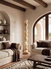 A modern bohemian-style living room featuring two cream-colored sofas with dark brown pillows and throws, a rustic wooden coffee table, and a tall, sculptural wooden floor sculpture in the center.