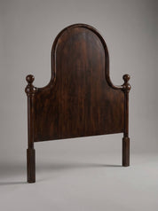 A dark brown wooden headboard with a tall arched backrest and turned postions at the sides, photographed against a neutral gray studio background.