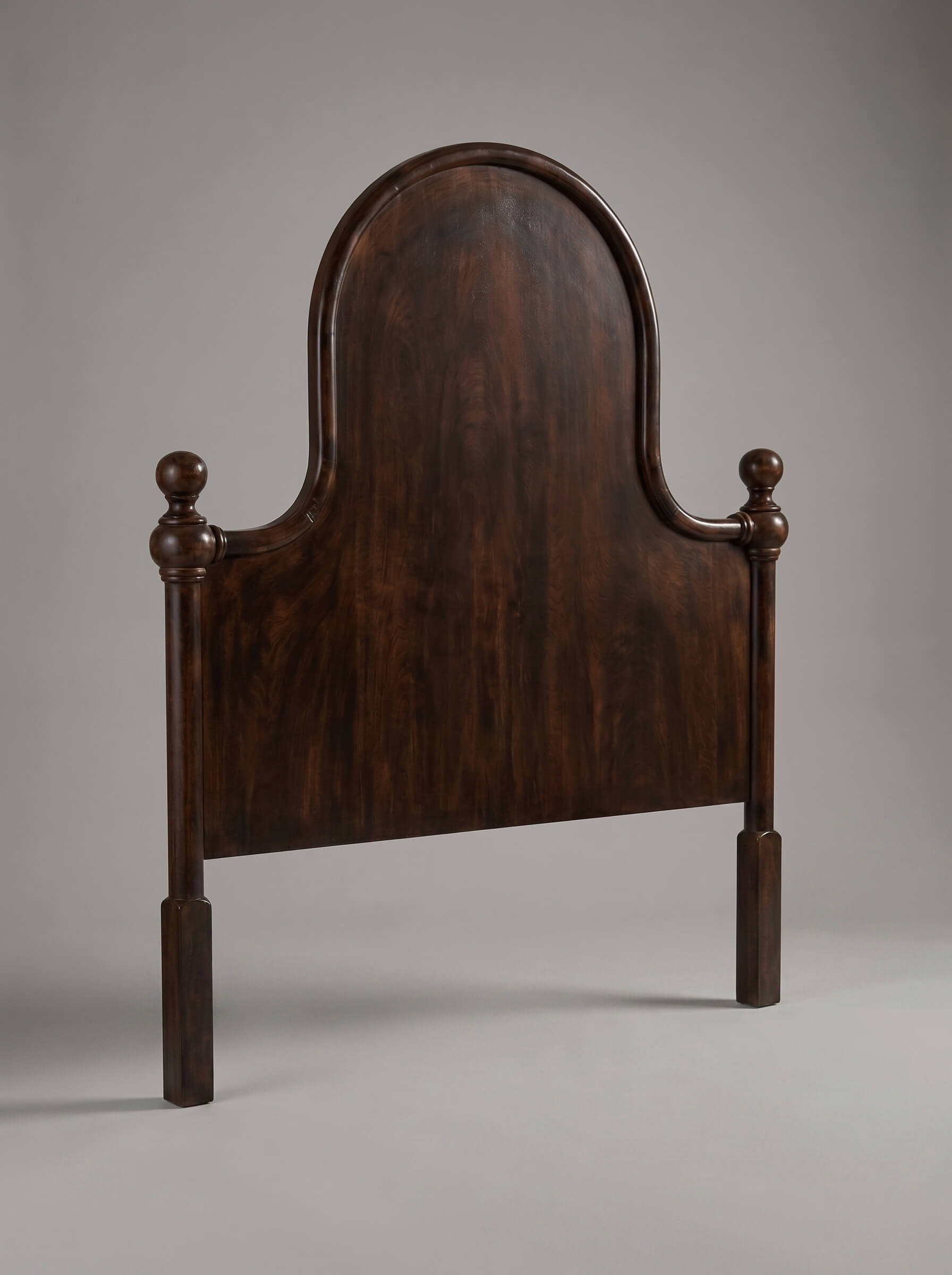 A dark brown wooden headboard with a tall arched backrest and turned postions at the sides, photographed against a neutral gray studio background.
