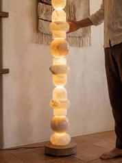A tall, illuminated stone floor lamp made of stacked glowing spheres on a round wooden base, shown in a rustic indoor room with a person's hand and lower body visible beside it.