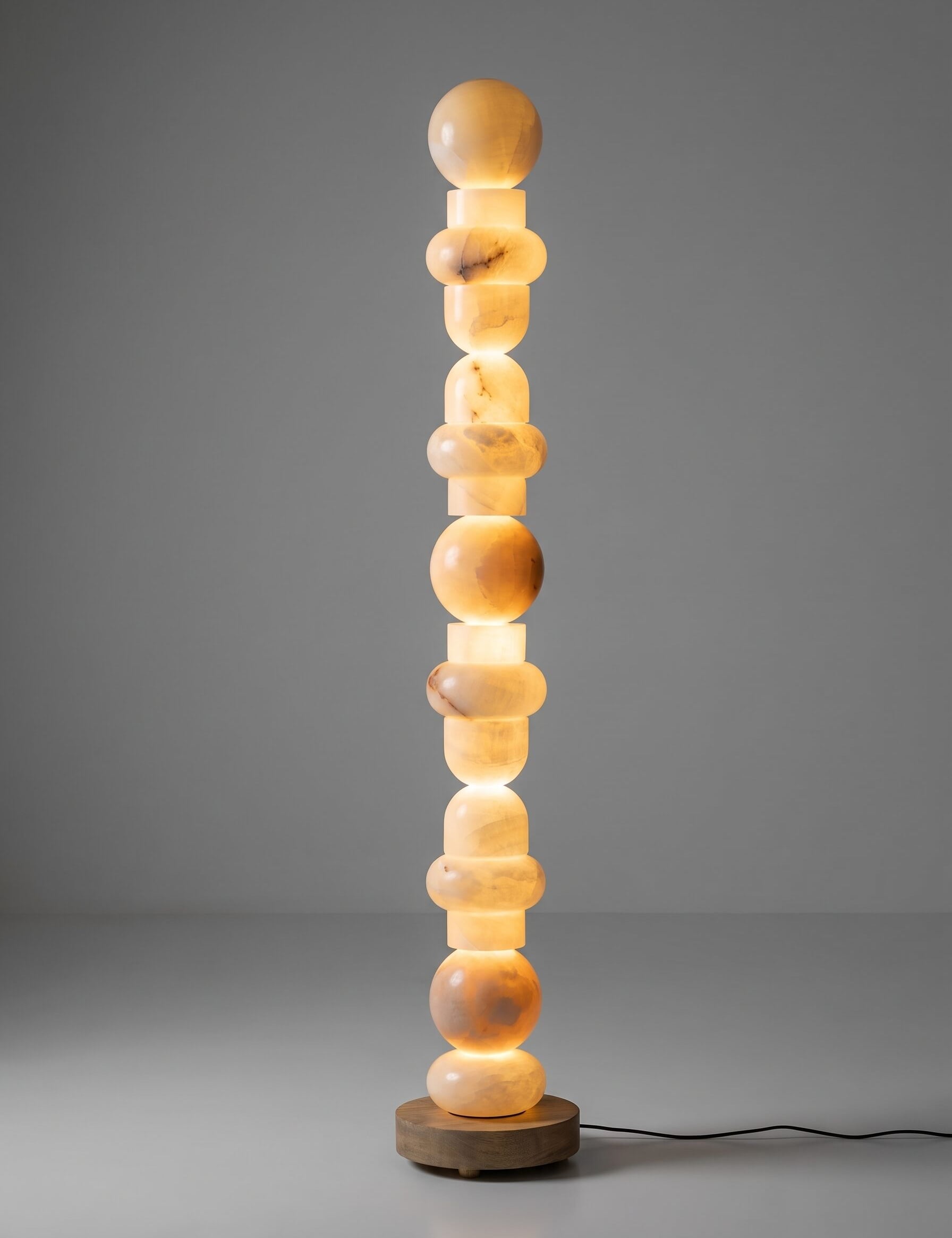 A modern sculptural floor lamp made of stacked glowing amber stone spheres on a round wooden base, photographed against a smooth gray studio background.