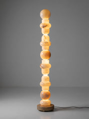 A modern sculptural floor lamp made of stacked glowing amber stone spheres on a round wooden base, photographed against a smooth gray studio background.