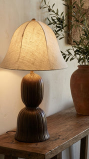 Table lamp with a textured base and linen shade on a wooden surface, with a plant in the background.