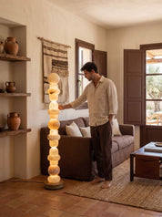 A man in a beige shirt and brown pants stands in a rustic living room next to a tall, illuminated wooden stacked floor lamp made of circular wood slices. The room features terracotta tile floors, a brown sofa, and wooden shelves with ceramic vases.