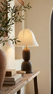 Table lamp with a white shade on a wooden table next to a plant and books.