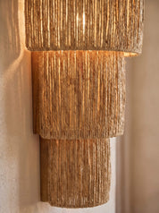 A tiered wall sconce light fixture made of natural woven jute or seagrass, illuminated with a warm glow against a textured beige wall.