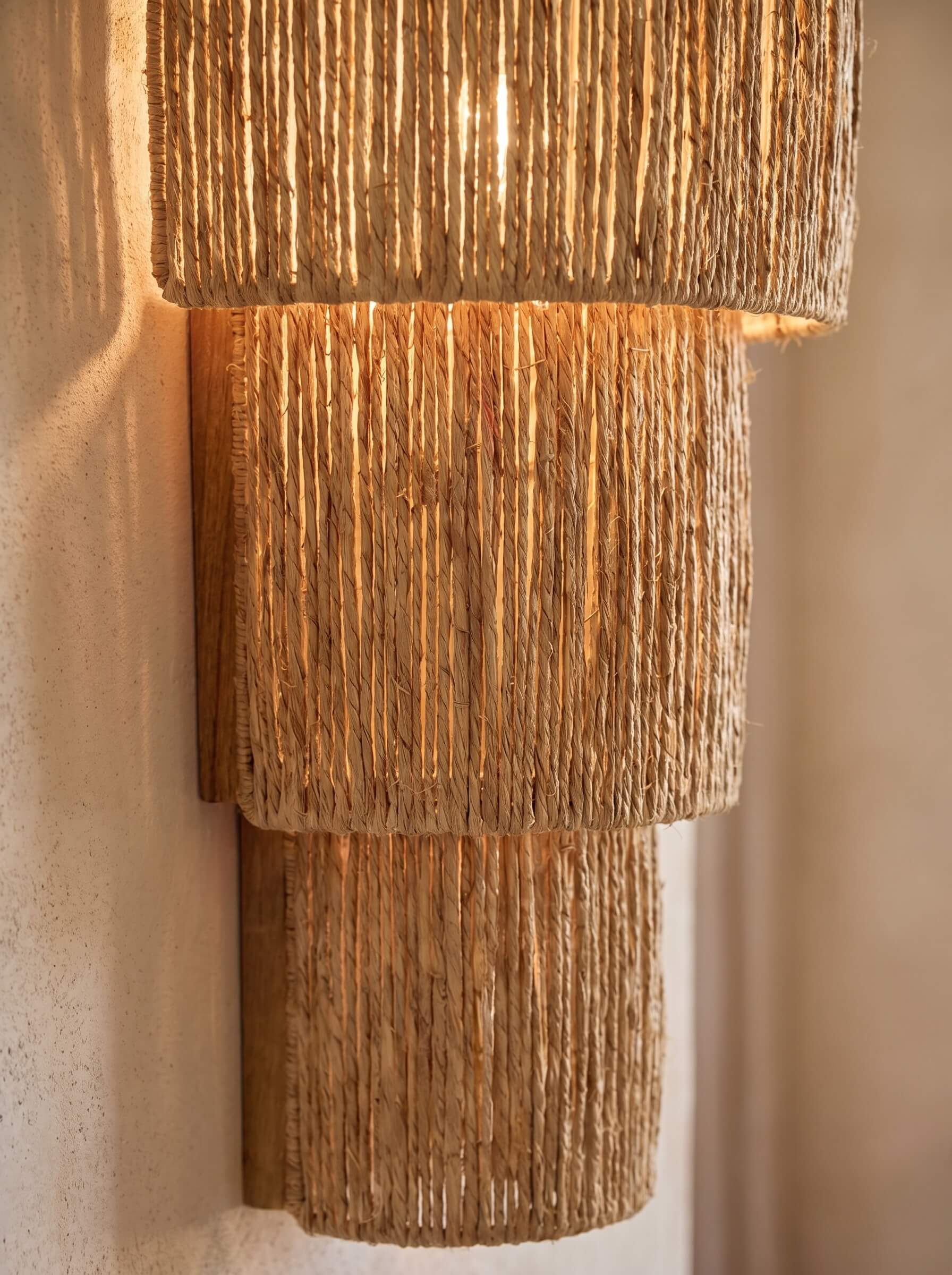 A tiered wall sconce light fixture made of natural woven jute or seagrass, illuminated with a warm glow against a textured beige wall.