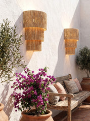 Two cylindrical wall sconces with woven natural-fiber shades mounted on a white stucco wall in a patio setting, with a wooden bench, pillows, and potted plants nearby.