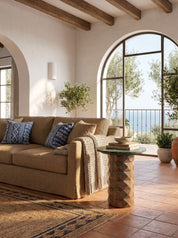 A styled interior scene showing a beige upholstered sofa with blue patterned pillows, a small round side table with a glass top, terracotta tile flooring, arched windows, and an outdoor view.