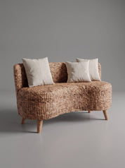 A small woven loveseat with a curved silhouette, upholstered in a textured natural fiber material, featuring three off-white throw pillows and tapered wooden legs, set against a solid gray studio background.