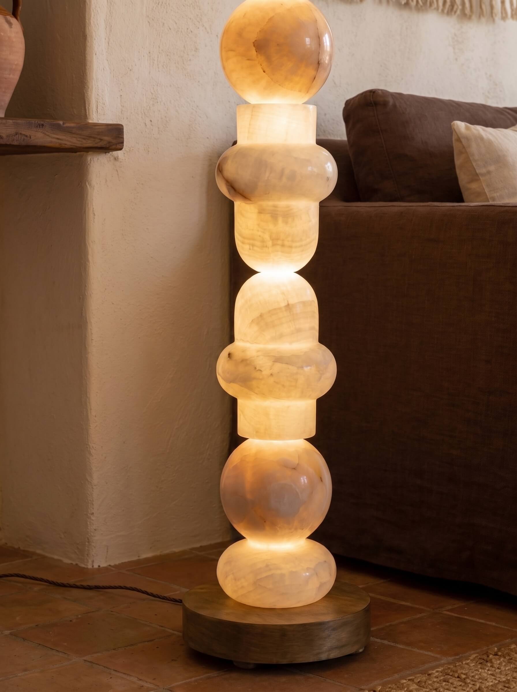 A modern sculptural floor lamp made of stacked, rounded translucent stone sections glowing with warm light, set on a round wooden base in a living room next to a brown sofa.