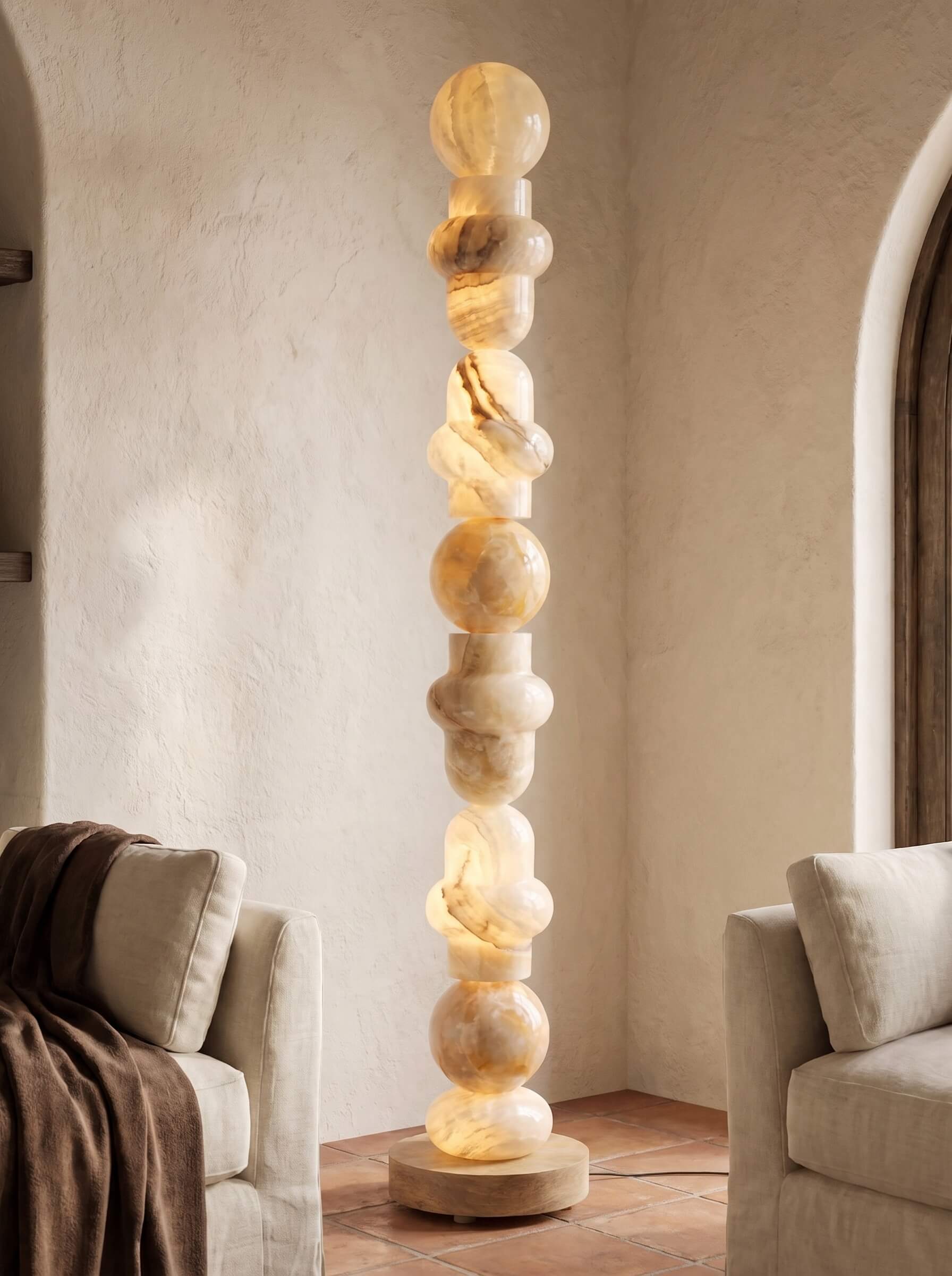 A tall, sculptural floor lamp made of stacked warm-toned stone spheres and twisted segments, glowing softly in a beige room with upholstered chairs and arches.