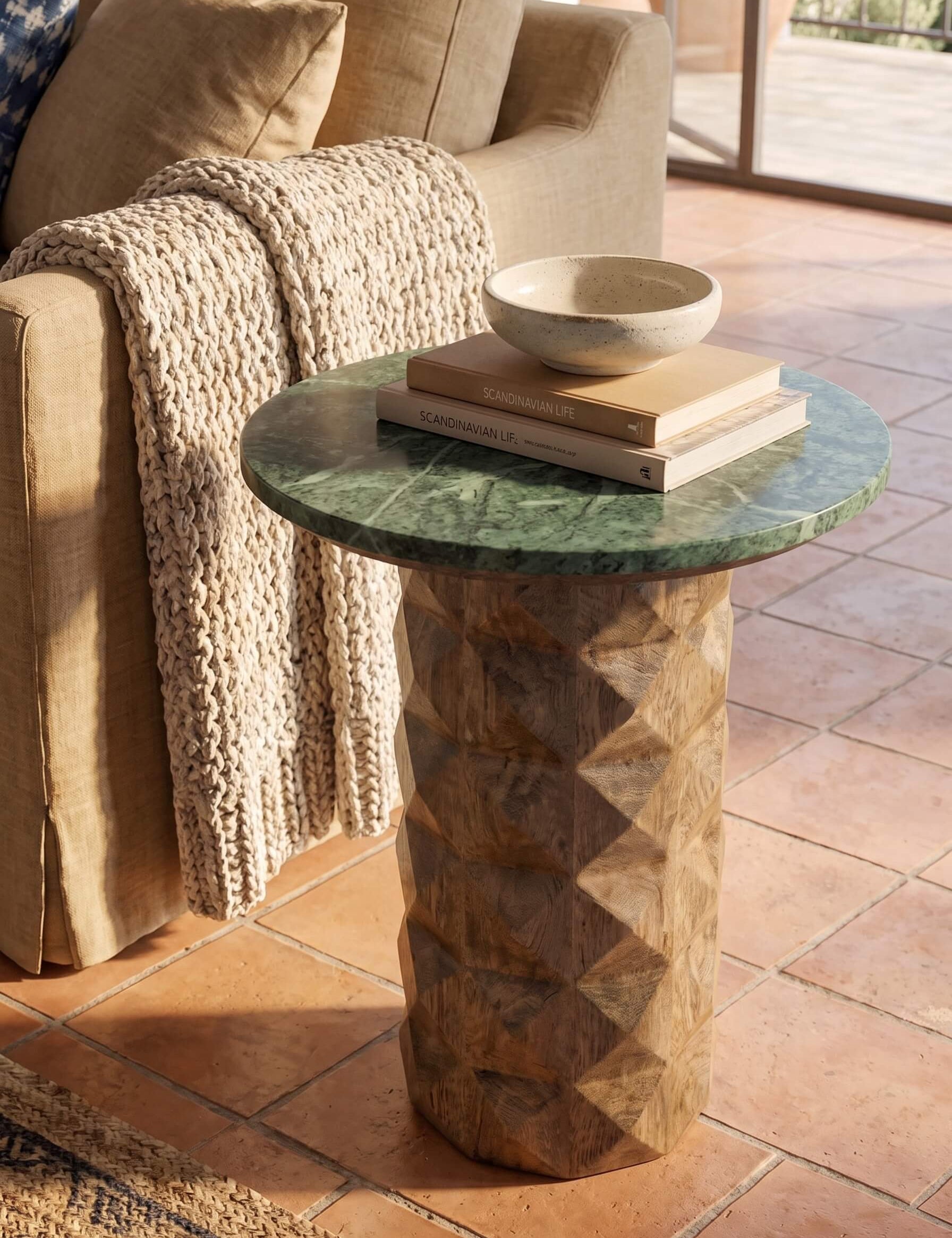 A round side table with a green marble top and a carved geometric wood pedestal base, styled in a warm living room setting next to a beige sofa with a chunky knit throw blanket.