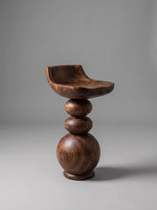 A sculptural wooden bar stool with a curved seat and stacked spherical legs, shown on a neutral gray studio background.