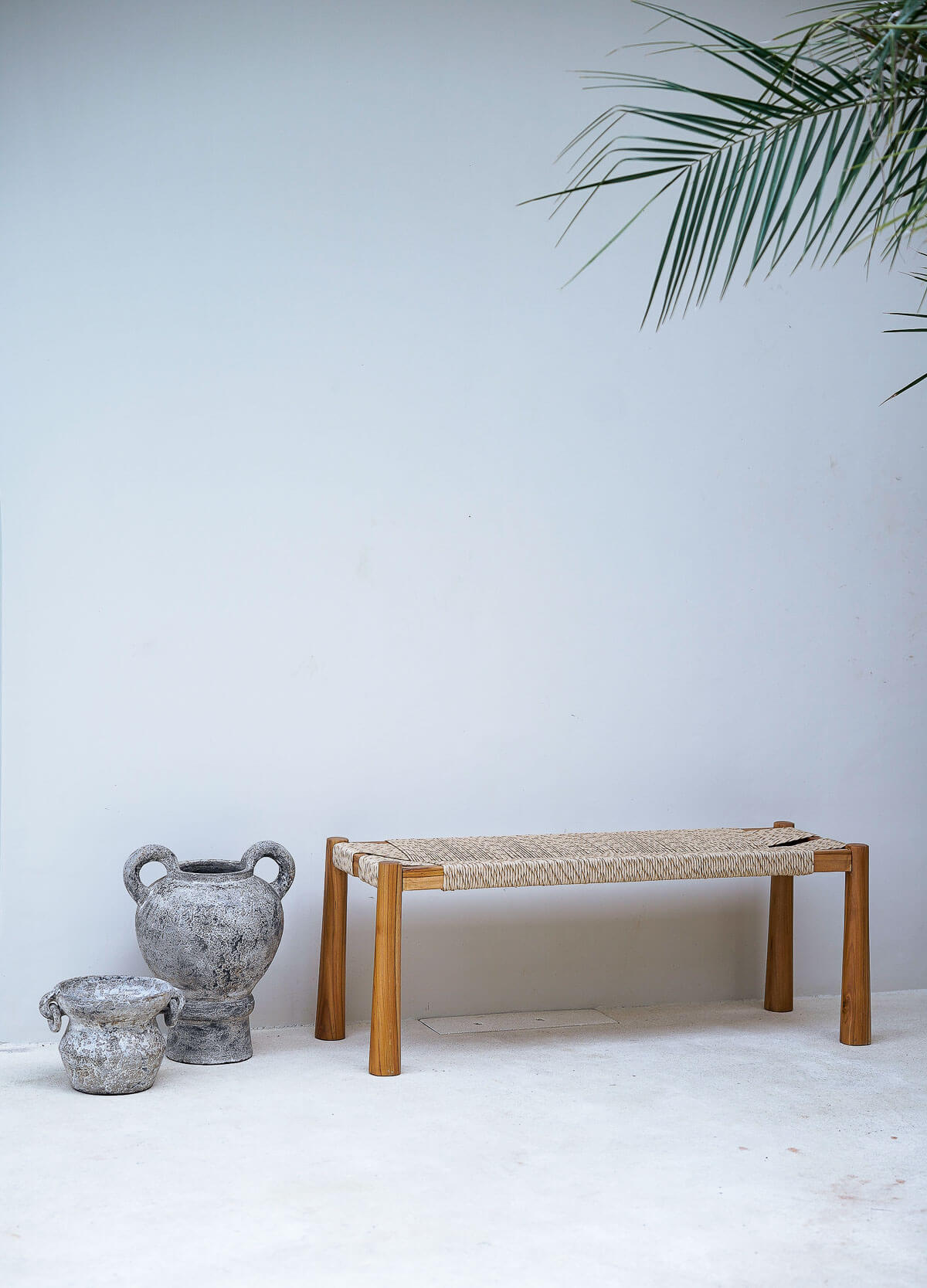 Wooden bench with woven seat against a white wall with decorative pots and a plant.