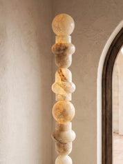 A tall, sculptural column lamp made of stacked organic-shaped polished stone sections in warm cream, beige, and tan tones, shown lit against a textured beige wall with part of a dark-framed arched doorway visible on the right.