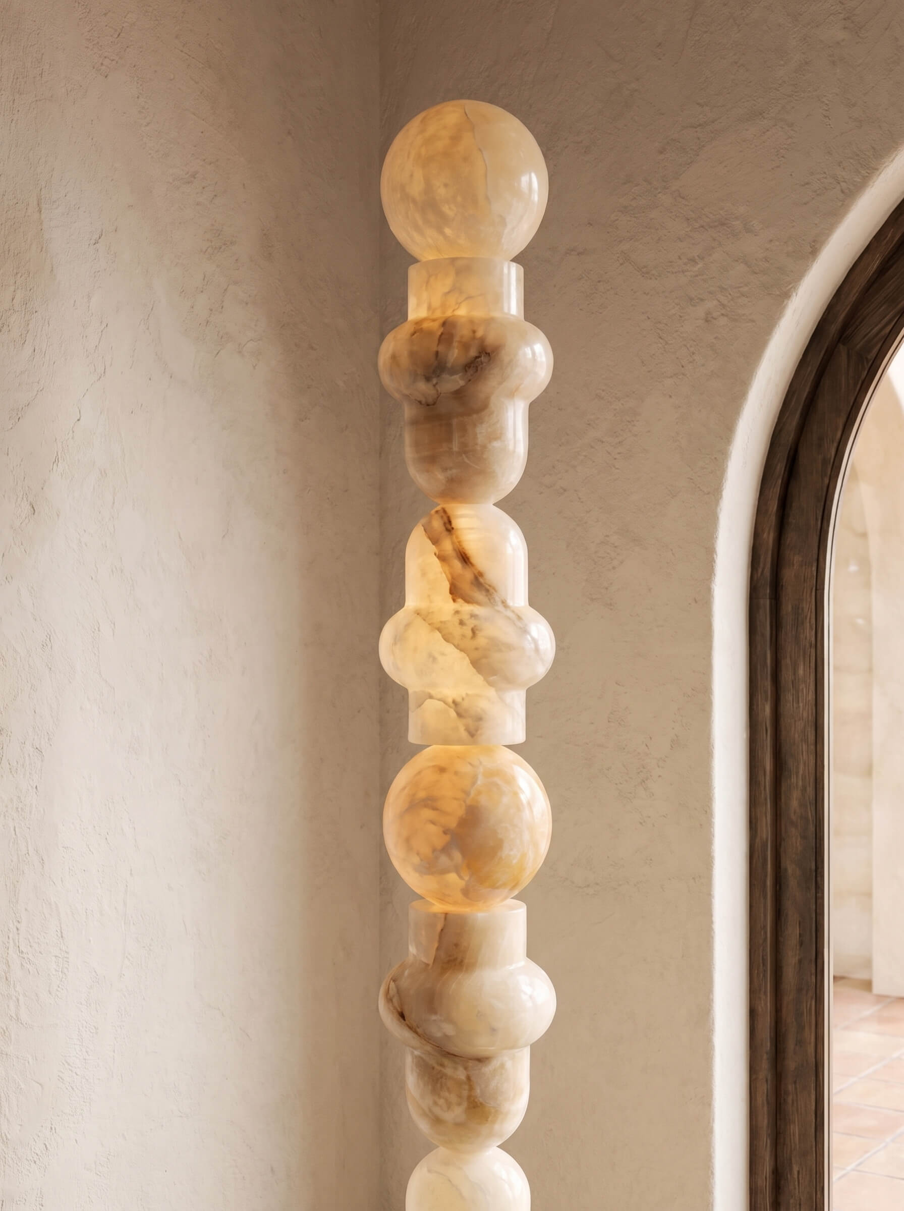 A tall, sculptural column lamp made of stacked organic-shaped polished stone sections in warm cream, beige, and tan tones, shown lit against a textured beige wall with part of a dark-framed arched doorway visible on the right.