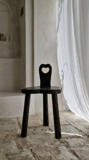 Black wooden chair with heart-shaped backrest in a room with white curtains and stone floor.