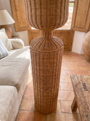 A tall, woven natural rattan or wicker floor lamp with a flared top and narrow neck, standing on a terracotta tile floor in a rustic living room next to a beige sofa.