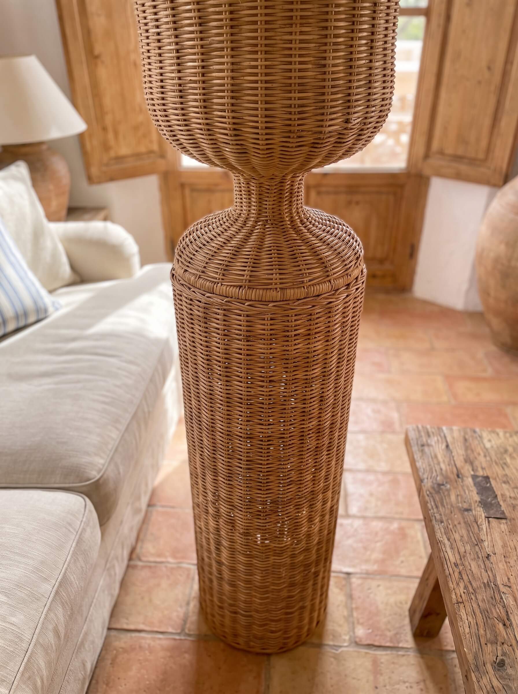 A tall, woven natural rattan or wicker floor lamp with a flared top and narrow neck, standing on a terracotta tile floor in a rustic living room next to a beige sofa.