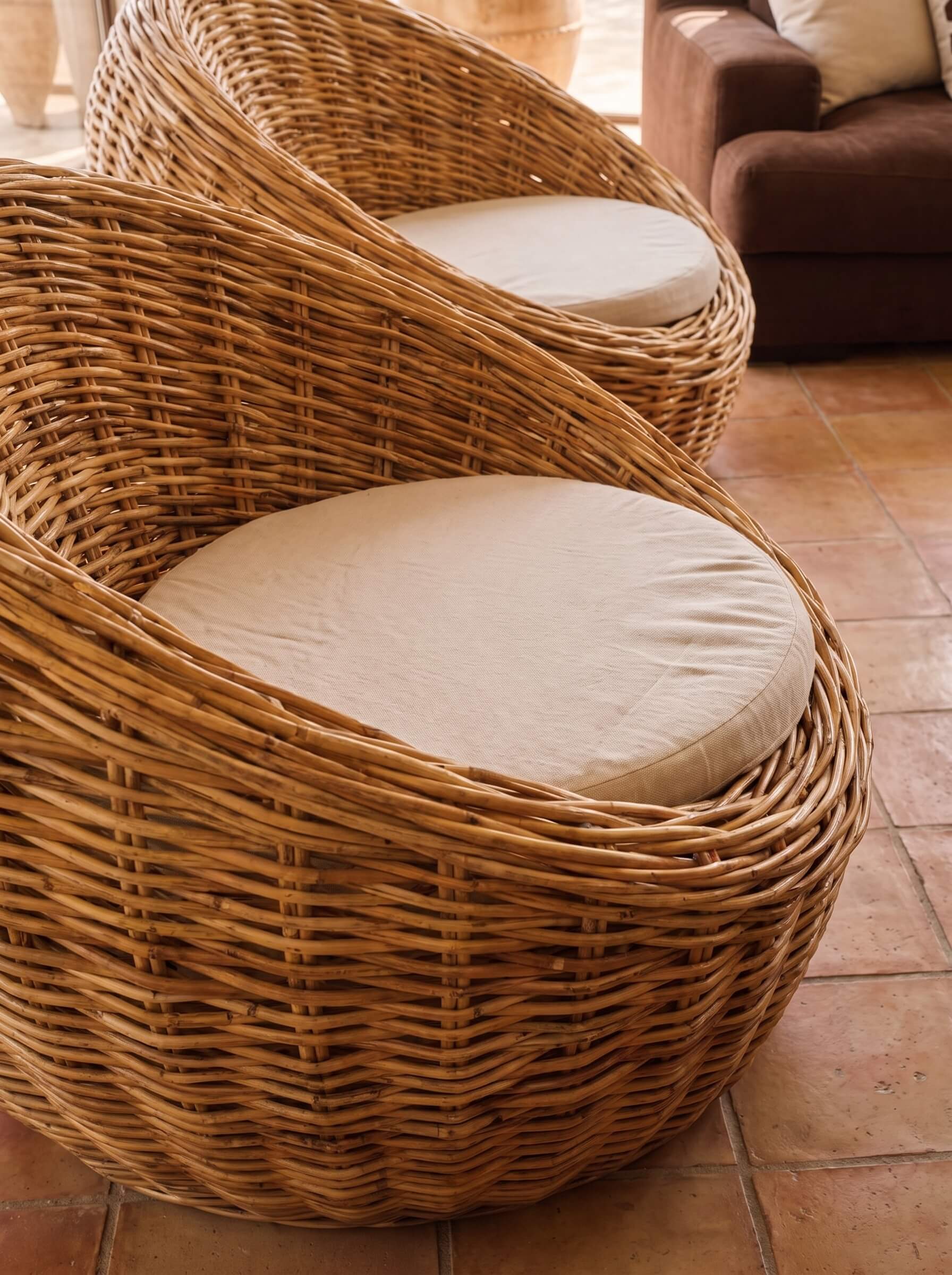 Two woven wicker lounge chairs with round off-white cushions, placed on terracotta tile flooring in a warm indoor setting.
