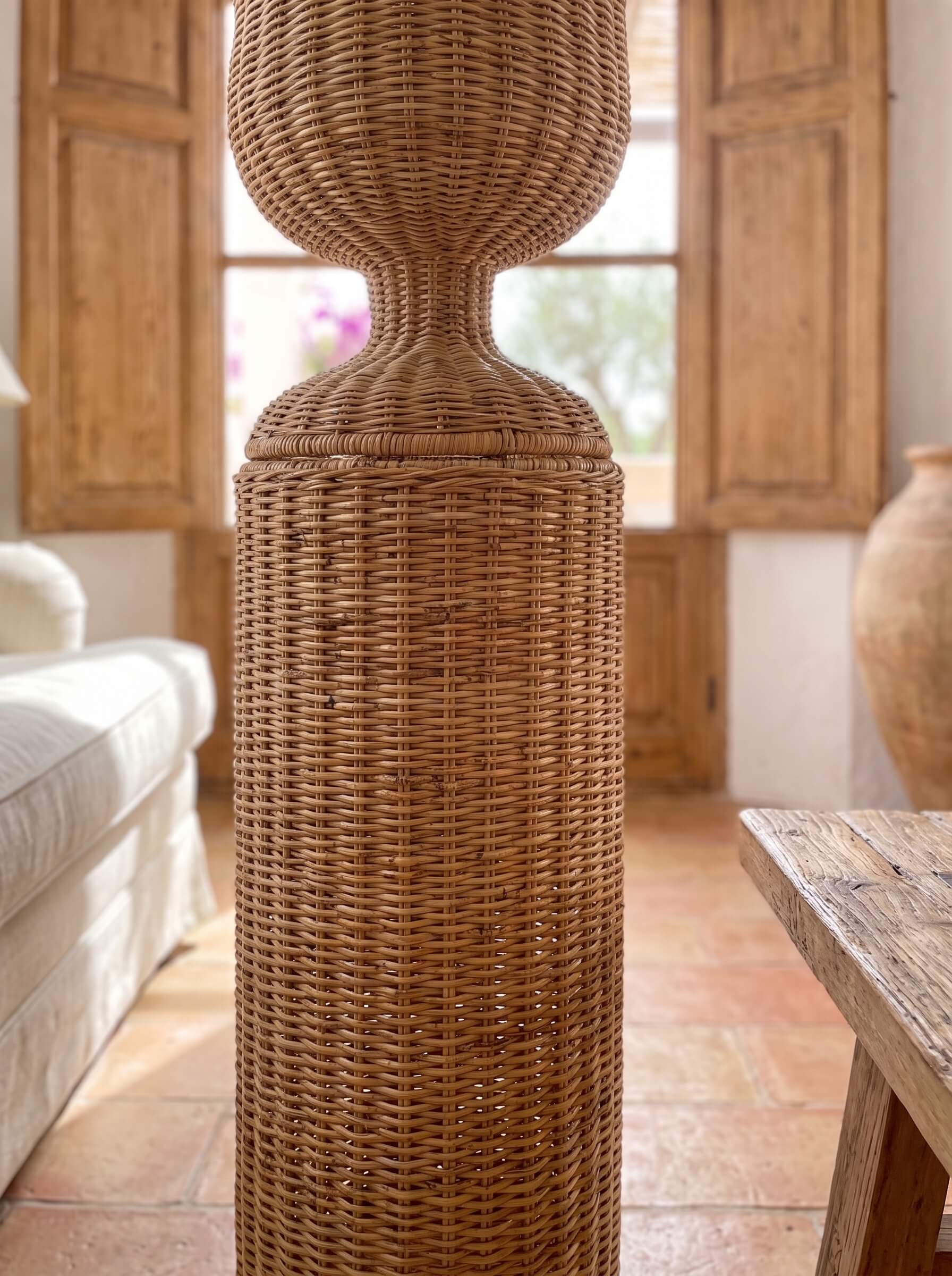 A tall, woven rattan decorative stand or vase with a flared top and cylindrical base, shown in a rustic indoor room with wood shutters, terracotta flooring, and a white sofa partially visible.