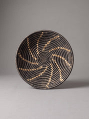 A round woven basket with a spiral pattern in black and natural tan, photographed against a neutral gray background.