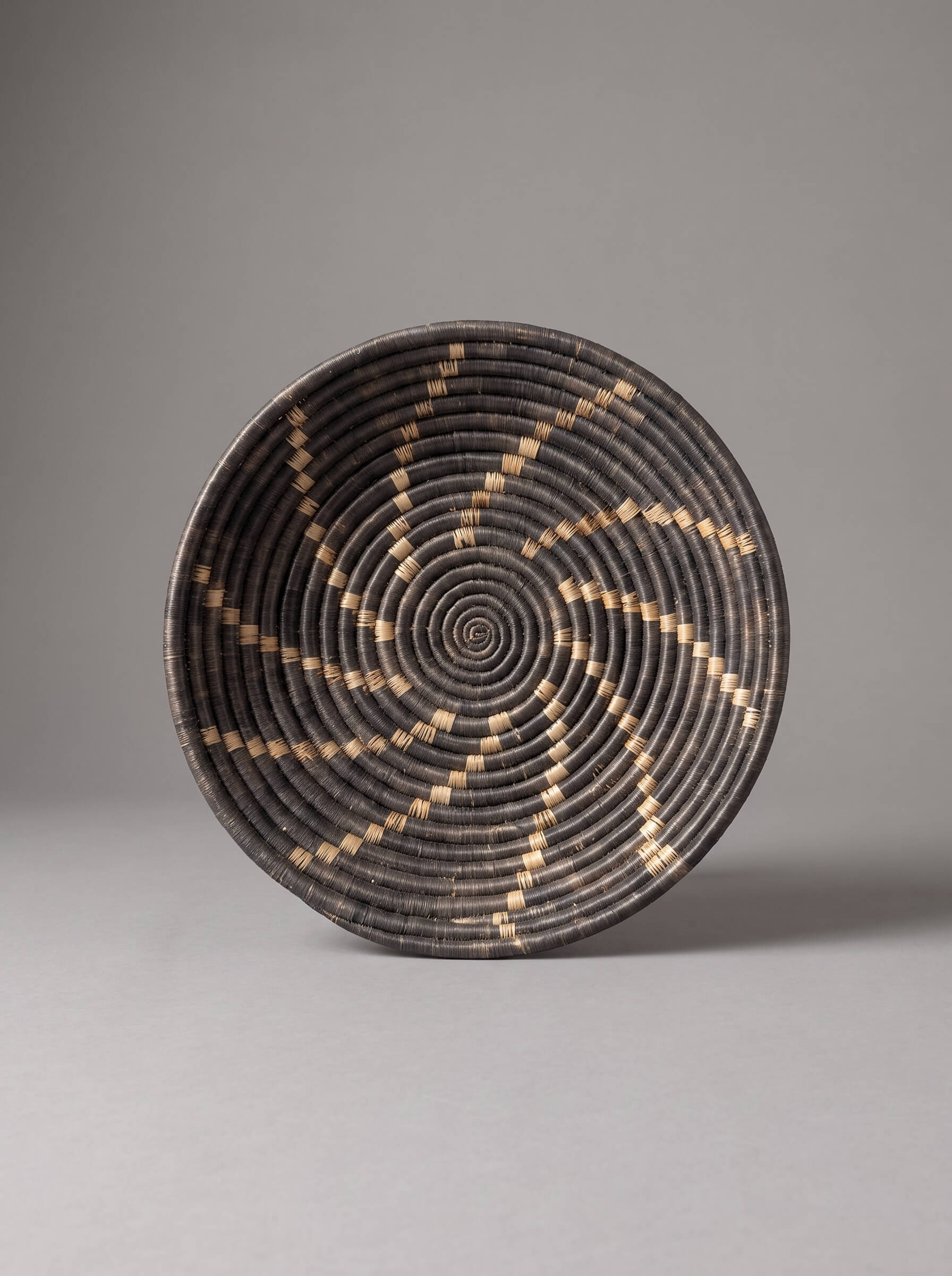 A round woven basket with a spiral pattern in black and natural tan, photographed against a neutral gray background.