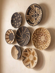 Seven hand-woven decorative baskets in various oval and round shapes, featuring intricate geometric patterns in black, brown, and natural beige, arranged on a textured beige wall.