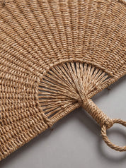 A close-up of a natural woven raffia or seagrass decorative fan, showing the detailed texture of the woven material and the handle loop on a neutral gray background.