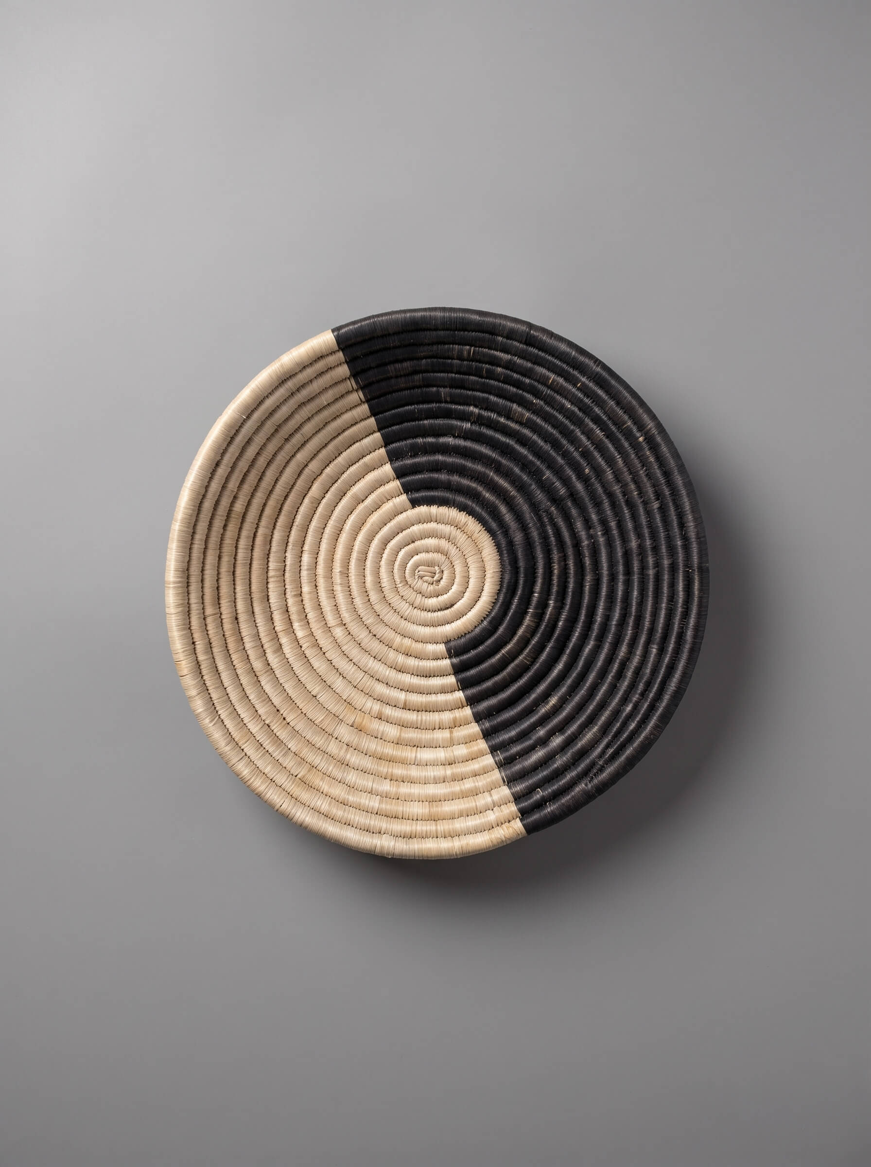 A round woven basket or tray with a half-and-half design, featuring natural beige on one side and black on the other, photographed from directly above on a neutral gray background.