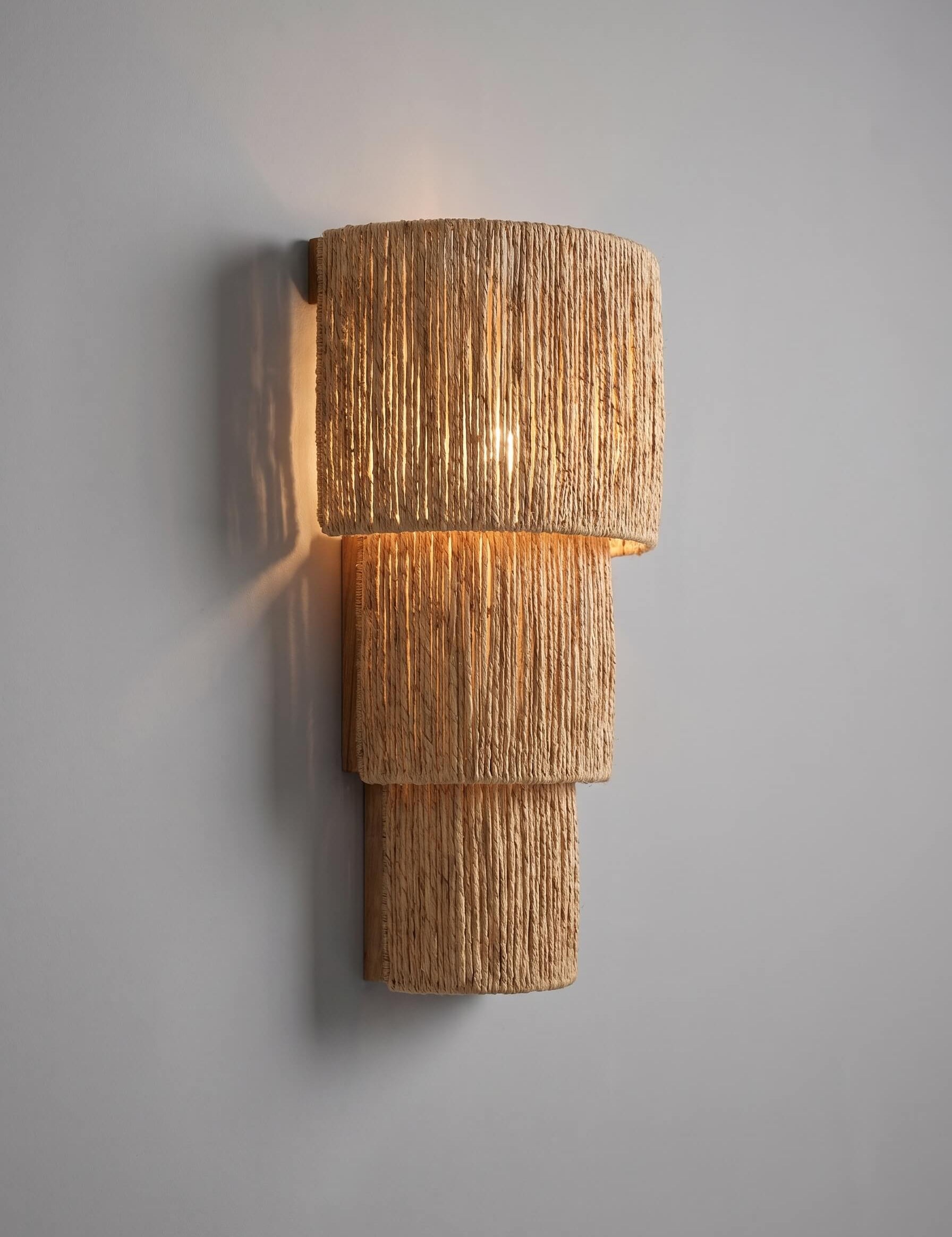 A modern tiered wall sconce with a textured, woven natural fiber shade, casting a warm golden glow against a neutral gray wall.
