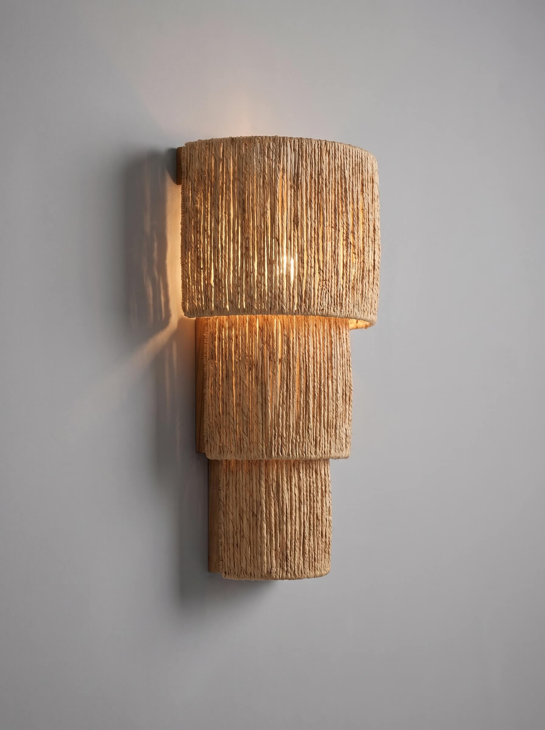 A modern tiered wall sconce with a textured, woven natural fiber shade, casting a warm golden glow against a neutral gray wall.