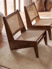 Two mid-century modern wooden lounge chairs with woven cane backrests and seats, placed on a beige rug in a warm, rustic interior setting.