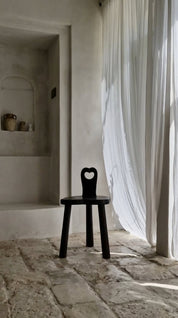 Black chair with a heart-shaped backrest in a room with stone flooring and white curtains.