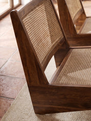 Close-up of a wooden lounge chair with woven cane backrest and seat, shown in a warm indoor setting with tiled floor and rug.