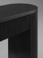 Close-up of a modern black wooden table with a curved top and vertically fluted side panels, photographed against a neutral gray background.
