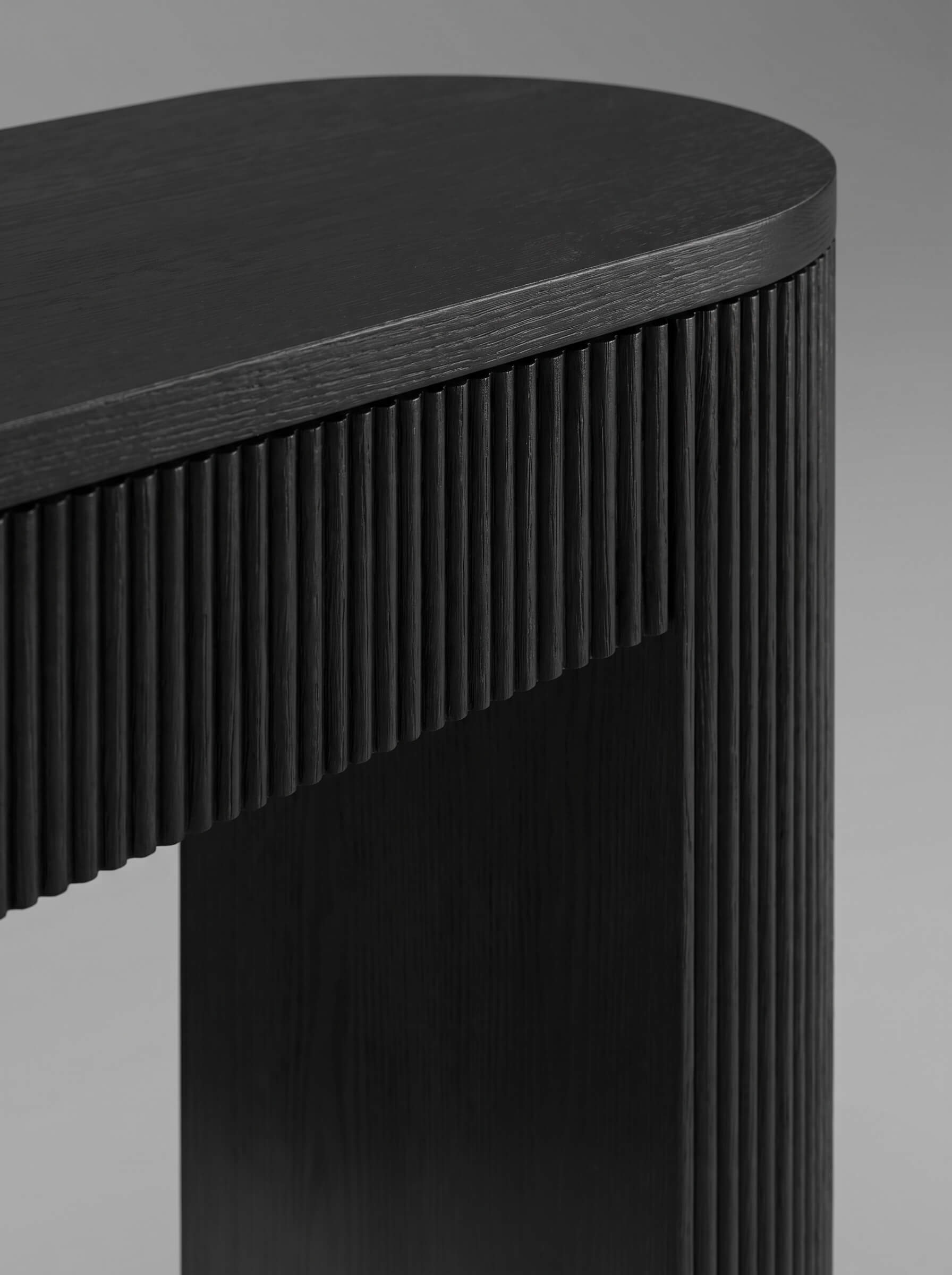 Close-up of a modern black wooden table with a curved top and vertically fluted side panels, photographed against a neutral gray background.